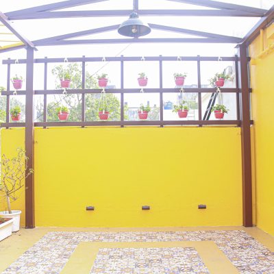 Vibrant yellow patio terrace featuring a patterned tiled floor, overhead metal trellis, and hanging potted plants.