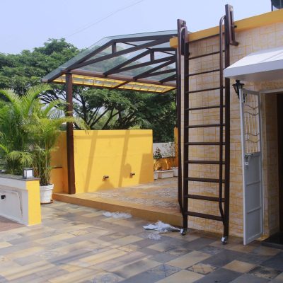 Rooftop patio terrace with yellow walls, a metal canopy structure, a fixed ladder, and potted palm plants.