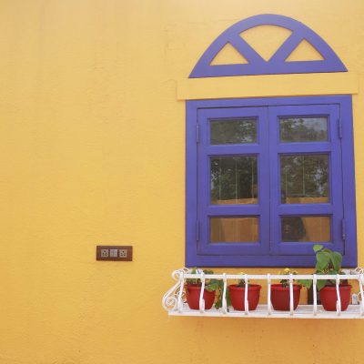 Vibrant yellow wall featuring a blue wooden window frame with an arched top and a white window box planter holding red pots.