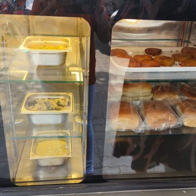 Glass display counter filled with stainless steel food trays, fried snacks, and burger buns.